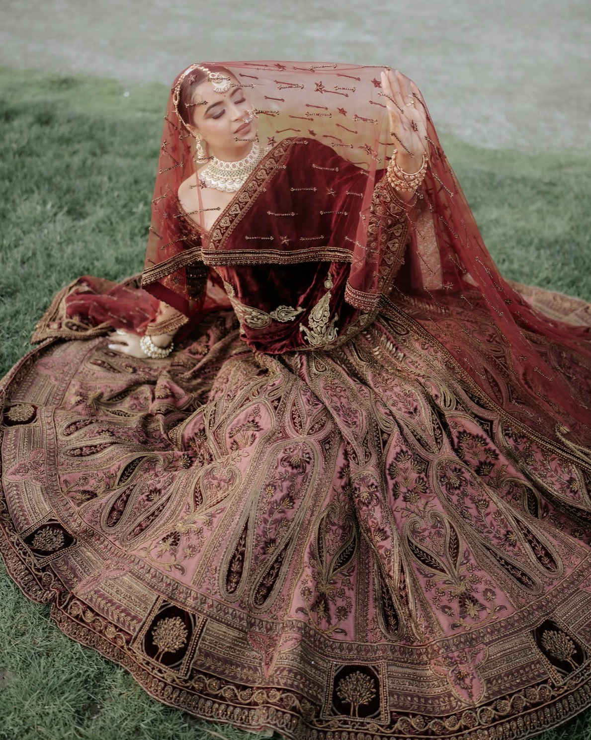 Bride seated on grass in a maroon velvet lehenga lifting her veil